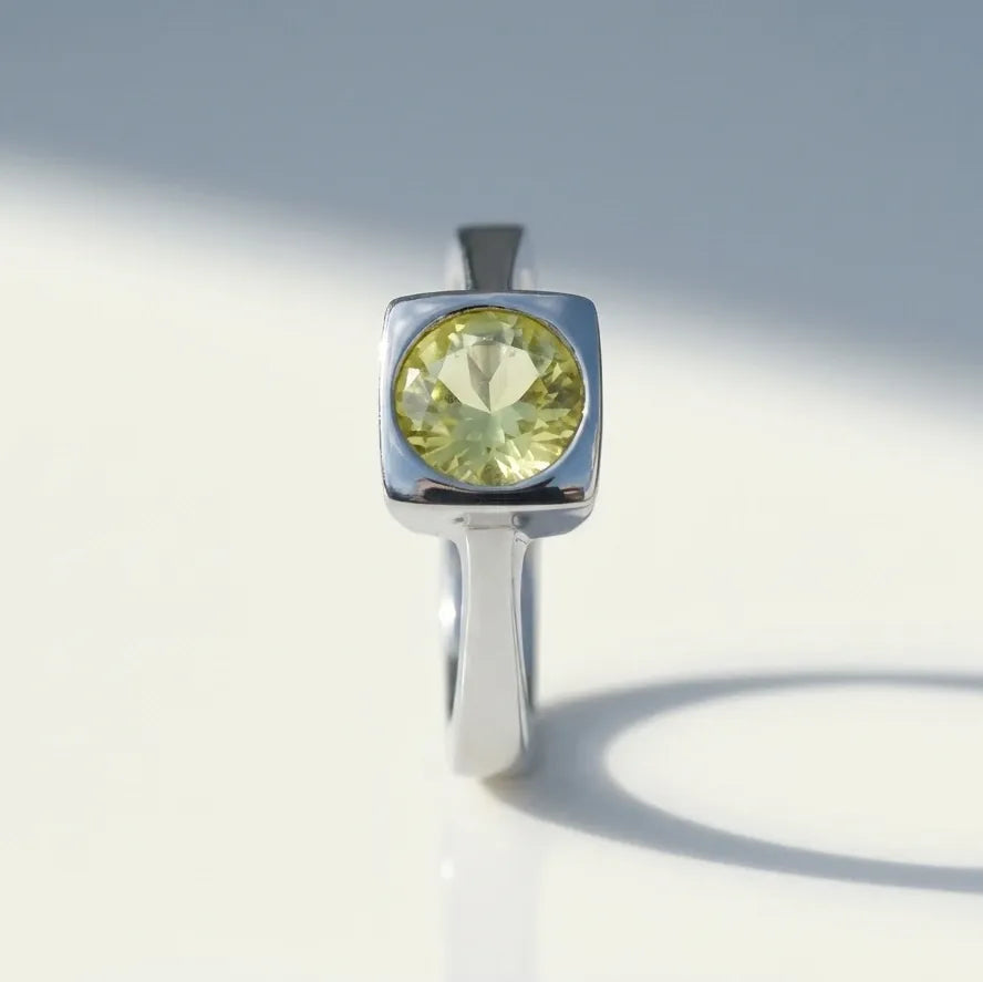 A silver ring with a square bezel setting holding a round faceted light yellow-green gemstone, photographed on a clean white surface with soft shadows.