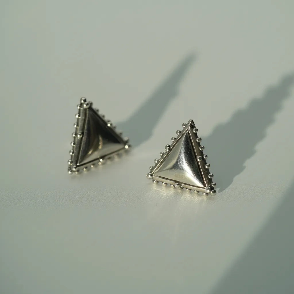 A pair of silver triangle stud earrings with beaded borders, resting on a light gray surface with soft shadows.