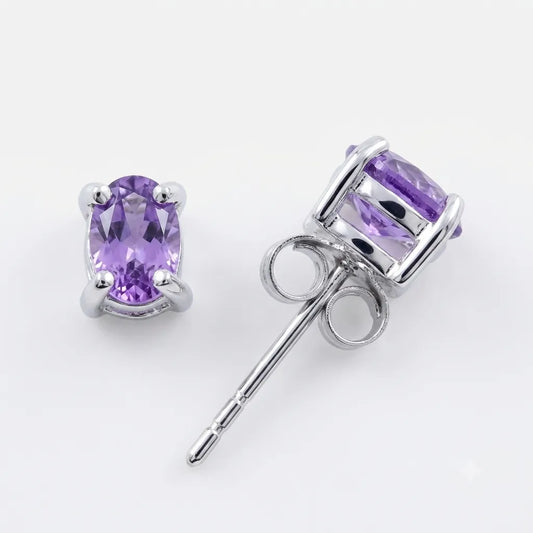 A pair of oval-cut purple amethyst stud earrings in silver-toned prong settings, with one earring facing forward and the other angled to show the post and backing, on a clean white background.