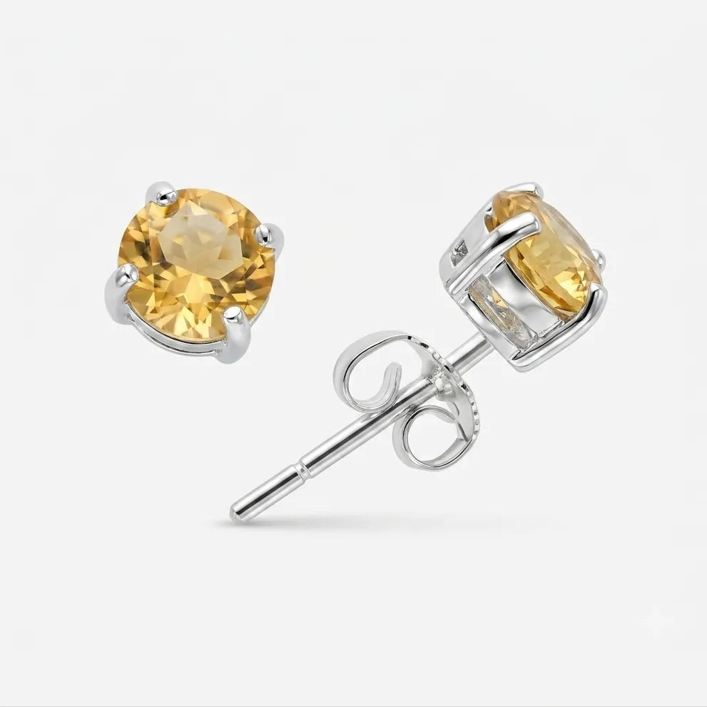 A pair of round-cut yellow gemstone stud earrings in silver prong settings, with one earring facing forward and the other angled to show the post and butterfly back, on a light gray background.
