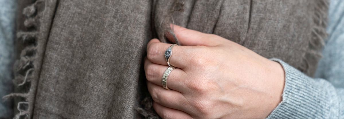 A close-up of a person wearing a soft taupe-brown fringed scarf over a light gray knit sweater, with one hand holding the scarf in place.