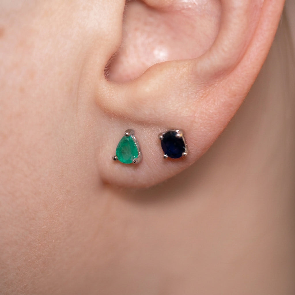 A close-up of a person's ear showing two small stud earrings: one light green teardrop-shaped stone in a silver setting and one dark navy blue oval stone in a silver setting.