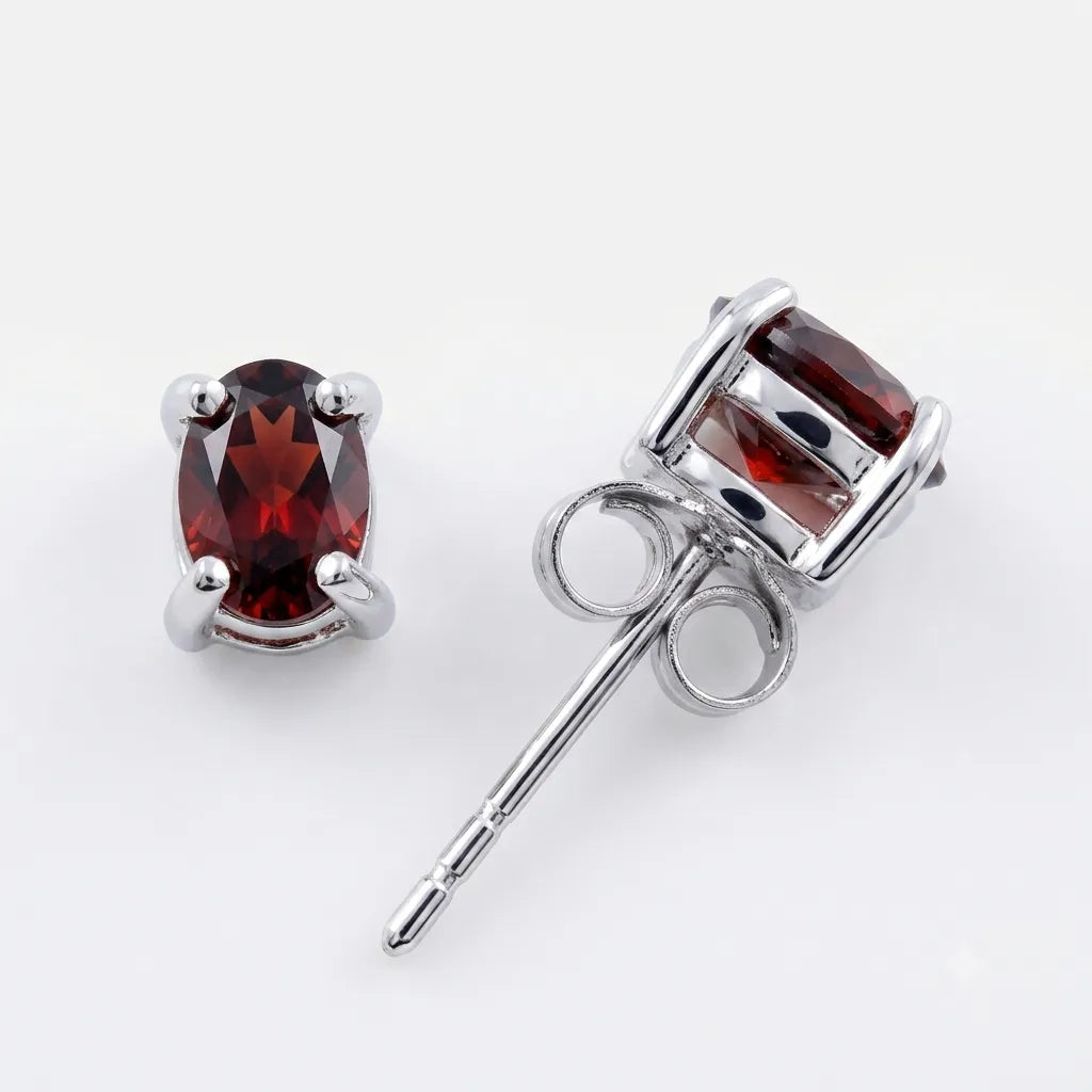 A pair of silver stud earrings featuring oval-cut deep red gemstones, one shown from the front and the other from the side/back to display the post and butterfly clasp, set against a plain white background.