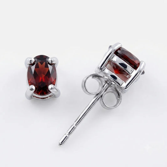 A pair of silver stud earrings featuring oval-cut deep red gemstones, one shown from the front and the other from the side/back to display the post and butterfly clasp, set against a plain white background.
