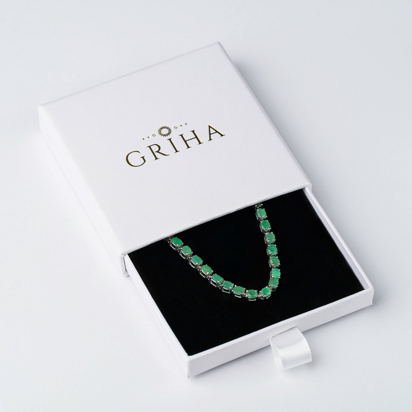 An emerald green gemstone bracelet displayed inside an open white GRIHA jewellery box with a black velvet insert, photographed on a clean white background.