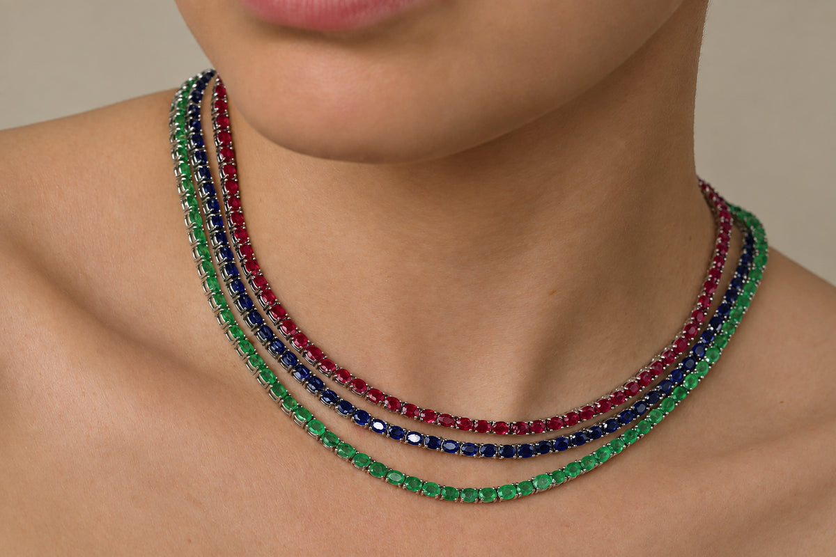 A close-up of a person's neck and upper chest wearing three layered tennis-style necklaces with faceted gemstones in emerald green, ruby red, and sapphire dark blue.
