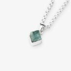 A silver chain necklace with a small, polished silver pendant holding a rough, faceted aquamarine green gemstone, shown on a light gray background.