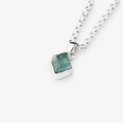 A silver chain necklace with a small, polished silver pendant holding a rough, faceted aquamarine green gemstone, shown on a light gray background.
