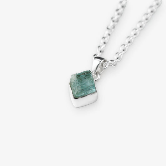 A silver chain necklace with a small, polished silver pendant holding a rough, faceted aquamarine green gemstone, shown on a light gray background.