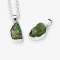 Two raw green peridot gemstone pendants in silver bezel settings, one hanging from a silver chain and one displayed flat, on a light gray background.