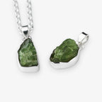 Two raw green peridot gemstone pendants in silver bezel settings, one hanging from a silver chain and one displayed flat, on a light gray background.
