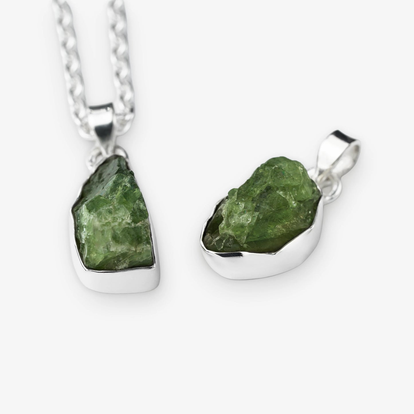 Two raw green peridot gemstone pendants in silver bezel settings, one hanging from a silver chain and one displayed flat, on a light gray background.