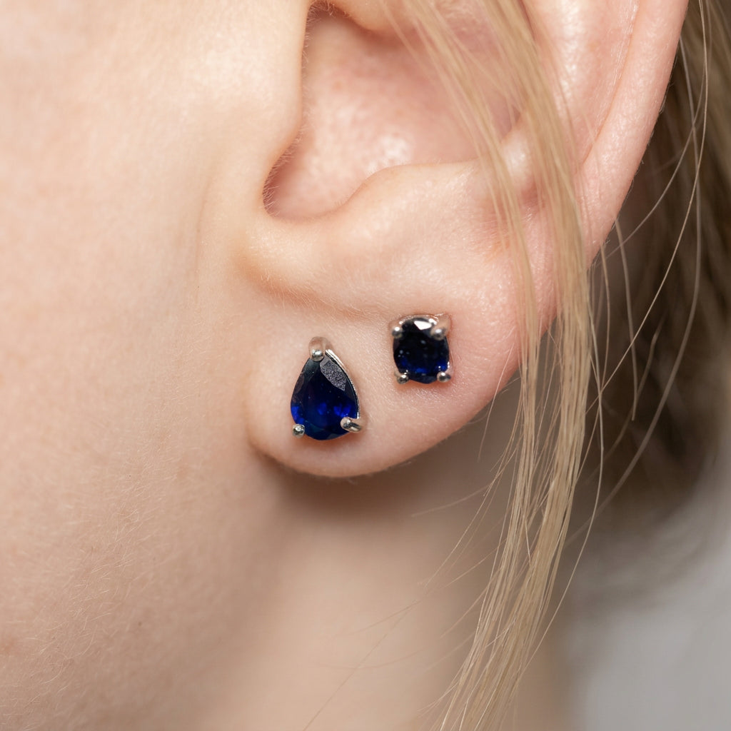 A close-up of a person's ear wearing two stud earrings: one teardrop-shaped dark blue gemstone earring and one oval dark blue gemstone earring in silver settings.