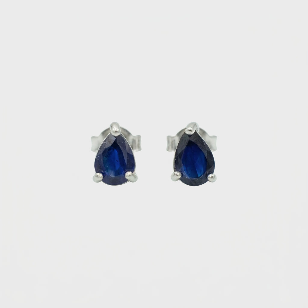 A pair of silver stud earrings featuring pear-cut deep blue gemstones in a three-prong setting, shown against a plain light gray background.