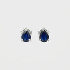 A pair of silver stud earrings featuring pear-cut deep blue gemstones in a three-prong setting, shown against a plain light gray background.