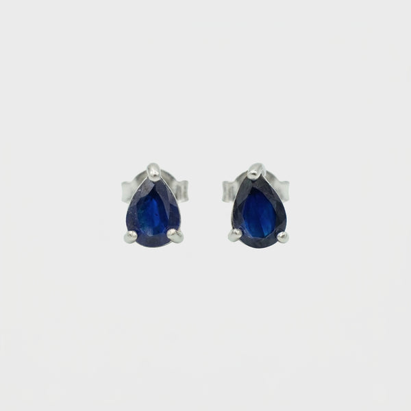 A pair of silver stud earrings featuring pear-cut deep blue gemstones in a three-prong setting, shown against a plain light gray background.