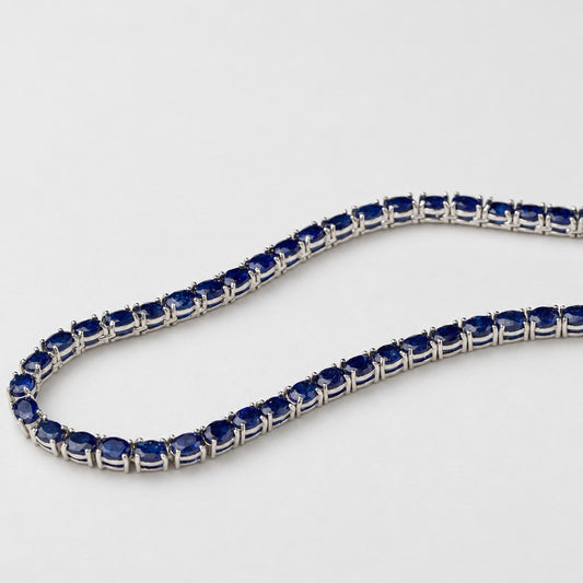 A tennis-style necklace with dark blue oval gemstones set in a silver-toned metal, photographed on a plain light background.
