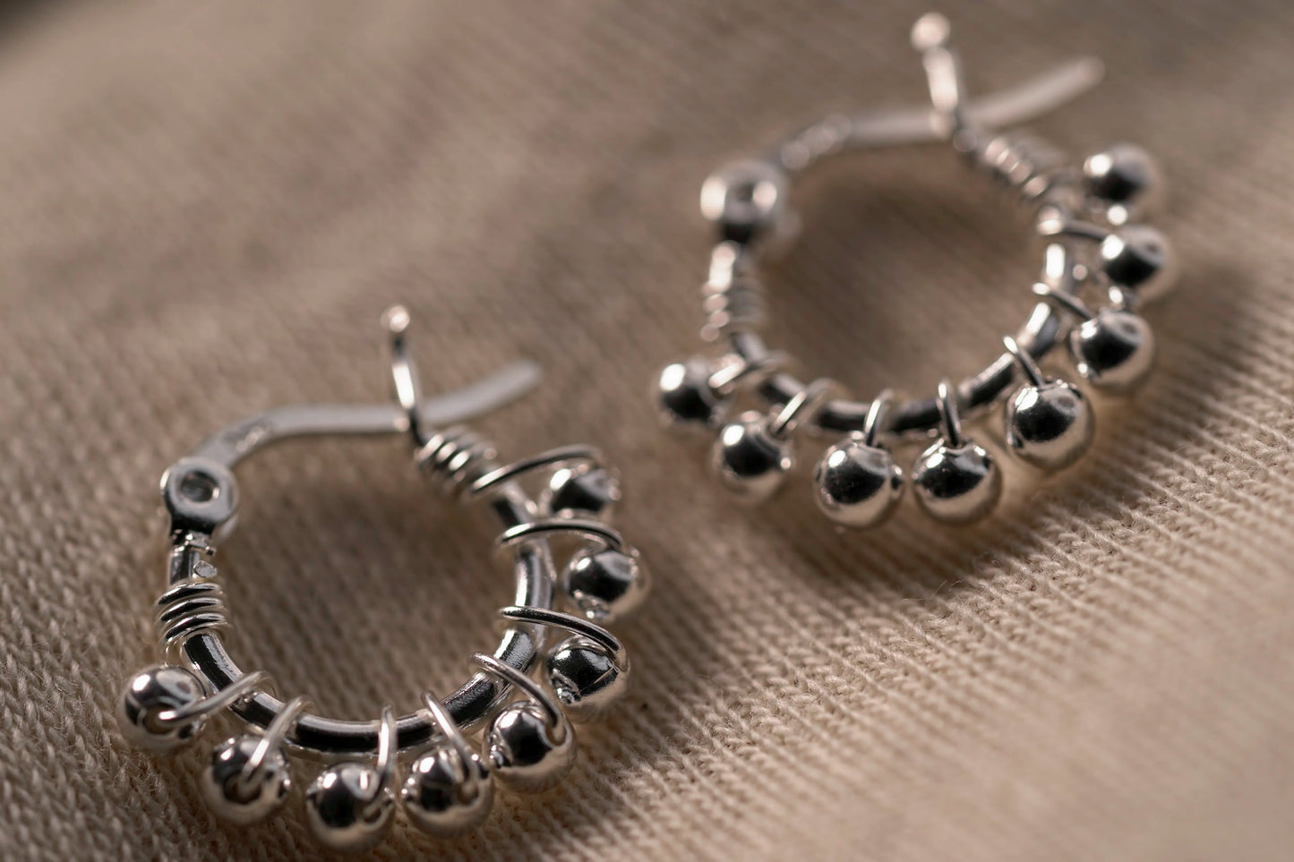 A pair of silver hoop earrings decorated with small metallic beads and wrapped wire details, displayed on a textured beige fabric background.