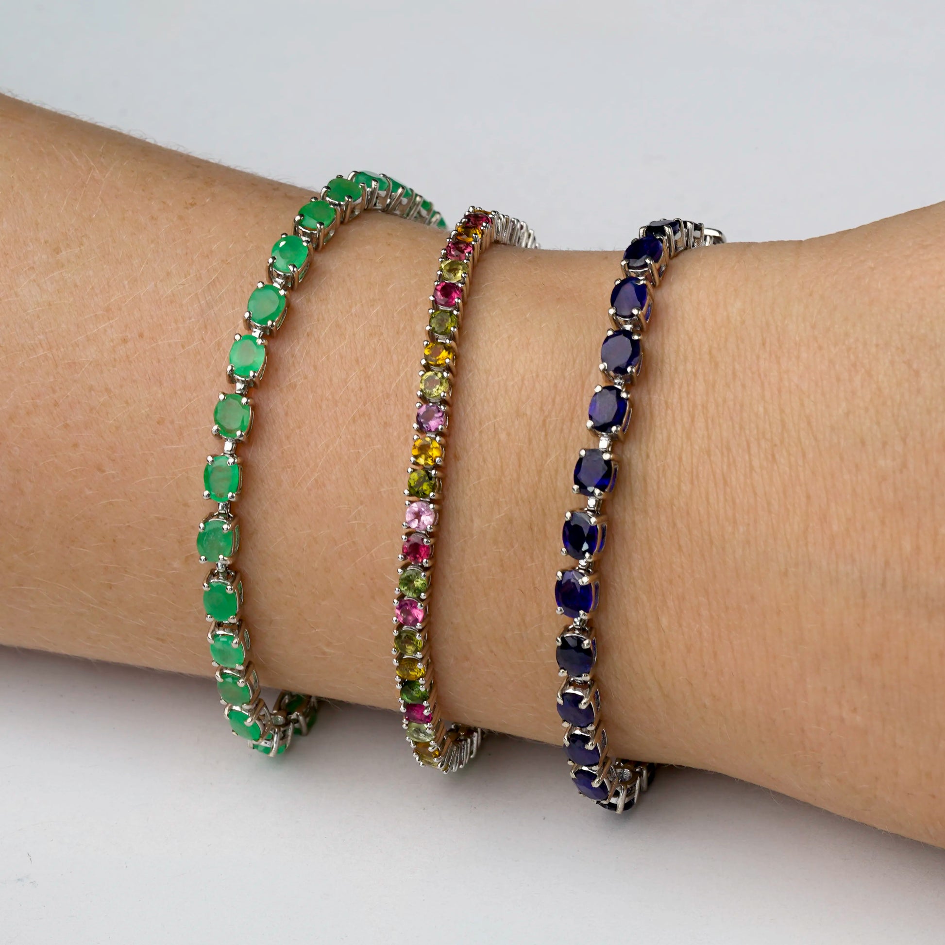 Three silver tennis bracelets with colourful gemstones are shown worn on a person's wrist. One bracelet features green emerald stones, the middle features tourmaline stones which are a mix of pink, yellow, and green stones, and the third features dark blue sapphire stones.