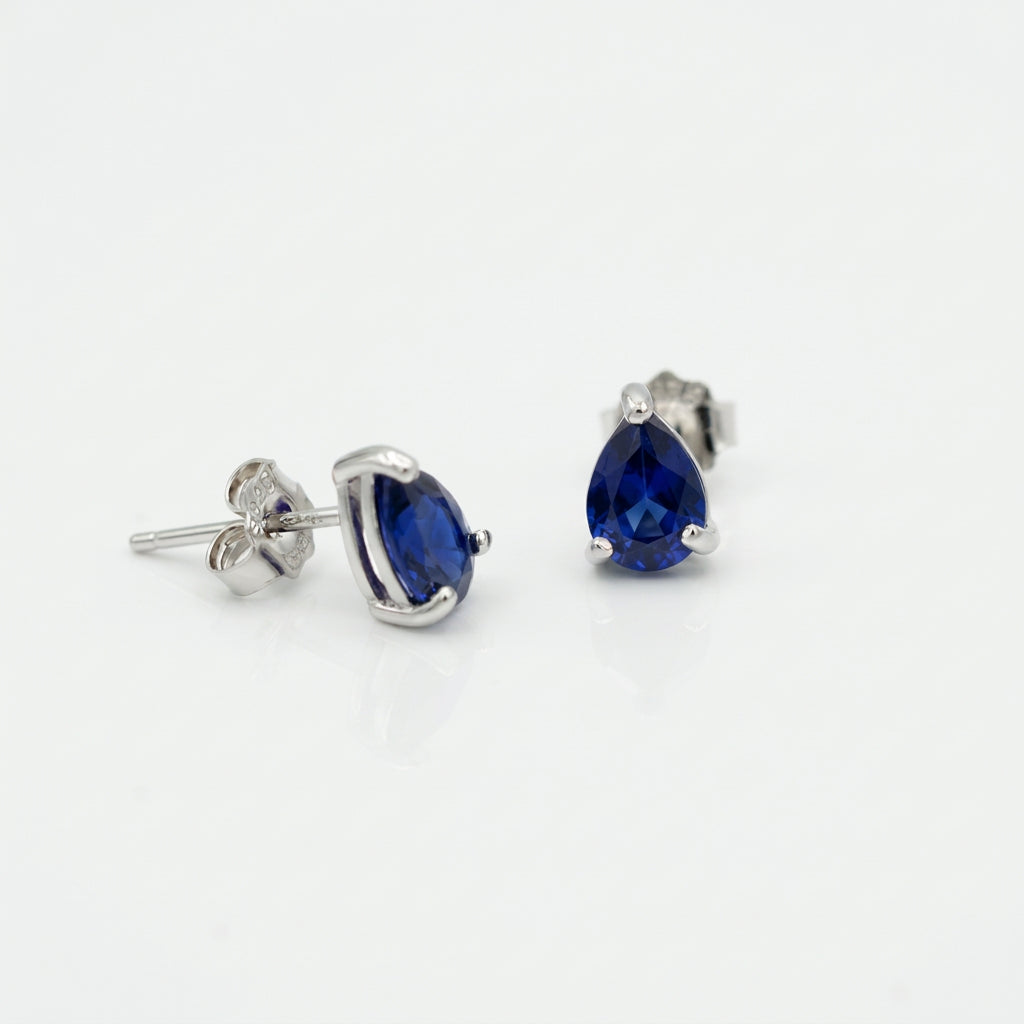 A pair of silver stud earrings featuring pear-cut deep blue gemstones, shown on a clean white background with one earring facing forward and the other angled to show the post and backing.