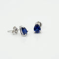 A pair of silver stud earrings featuring pear-cut deep blue gemstones, shown on a clean white background with one earring facing forward and the other angled to show the post and backing.