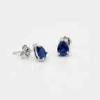 A pair of silver stud earrings featuring pear-cut deep blue gemstones, shown on a clean white background with one earring facing forward and the other angled to show the post and backing.