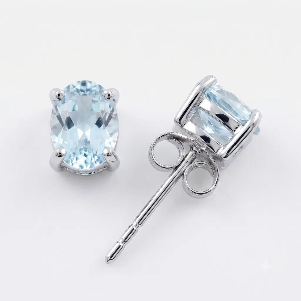 A pair of oval-cut light blue gemstone stud earrings in silver-toned metal settings, with one earring facing forward and the other angled to show the post and backing, on a clean white background.