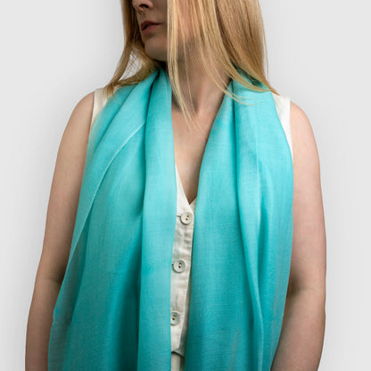 A woman wearing a lightweight turquoise blue scarf draped over her shoulders, paired with a white sleeveless top. The photo is cropped from the mouth down against a clean, light gray background.