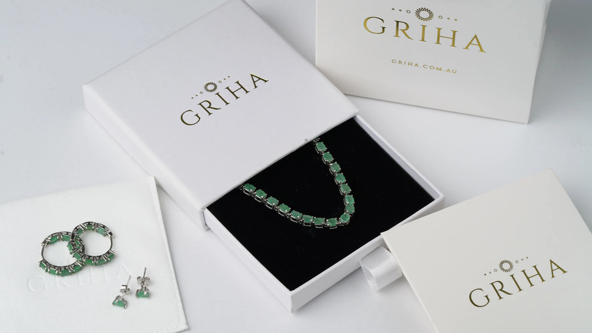 A jewelry set from the brand Griha, featuring a green stone necklace in a black-lined presentation box, matching green stone hoop earrings, and stud earrings, arranged on a white surface with white branded boxes.