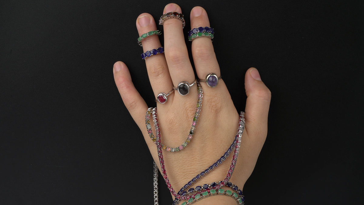 A hand wearing multiple colorful gemstone rings and matching beaded bracelets against a solid black background.