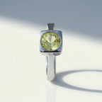 A silver ring with a square bezel setting holding a round faceted light yellow-green gemstone, photographed on a clean white surface with soft shadows.